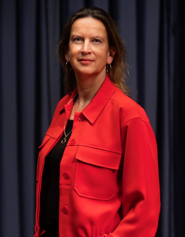 Medium-close portretfoto van Emily Ansenk, ze staat met haar handen in haar zakken en kijkt recht de camera in. Ze draagt een rood jack.