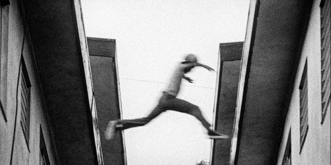 In a grainy black-and-white film still full of movement shot looking up at a roof from a small alleyway, a young male figure leaps from one roof to the next.