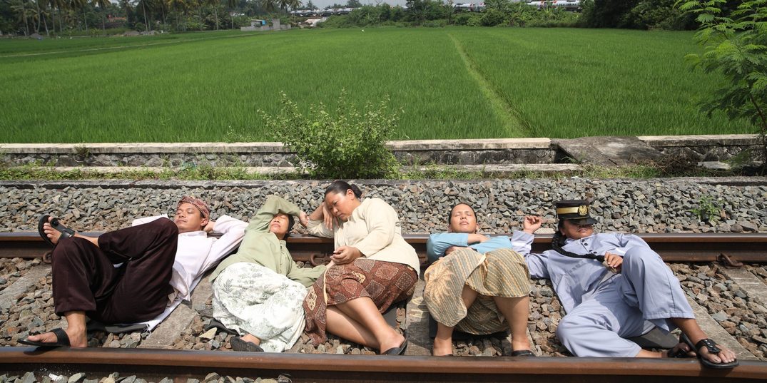 In a lush green landscape, in the foreground, five people lie napping on train tracks