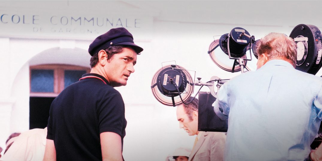 Regisseur Jacques Demy, een jonge, slanke man met donker haar, een donkerblauwe polo en een kapiteinspetje, staat met zijn rug naar de camera op een drukke filmset. Naast hem zit een operateur van een filmcamera op een verhoogd krukje.