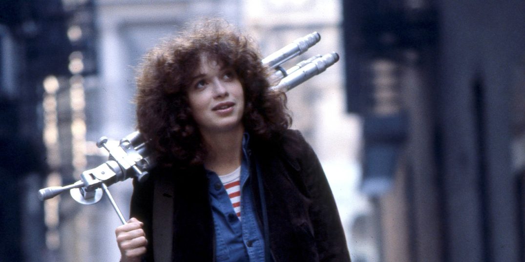 In a film still from the 1978 film Girlfriends, a smiling young woman with curly hair walks in downtown New York, a photo camera around her neck and carrying a tripod on her shoulder