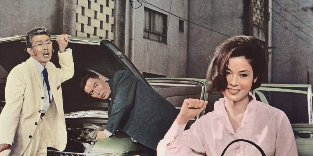 Op een ingekleurde foto uit 1962 steekt een vrouw in een roze blouse haar rechtervuist in de lucht – ogenschijnlijk als teken van overwinning – met een brede glimlach op haar gezicht. Vlak boven haar rechterschouder kijken twee mannen haar na, terwijl ze over de open motorkap van een auto leunen.