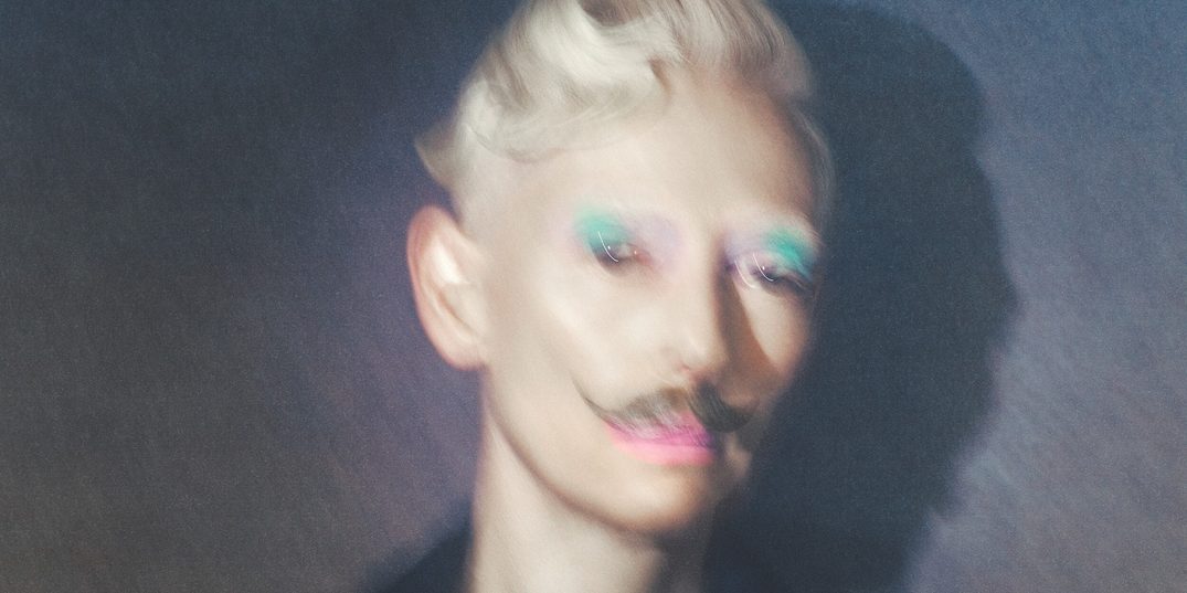 In a slightly blurry photo, Tilda Swinton stands against a mottled grey background, wearing a dark double-breasted suit. Her short blonde hair is curled up into a coiled quiff, she wears a popping turqouise eyeshadow, a light pink lipstick, and a curling brown moustache.