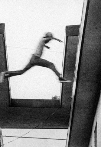 In a grainy black-and-white film still full of movement shot looking up at a roof from a small alleyway, a young male figure leaps from one roof to the next.