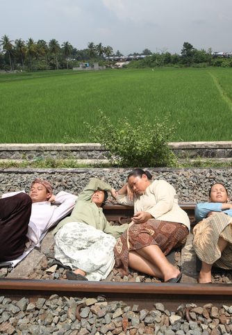 In a lush green landscape, in the foreground, five people lie napping on train tracks