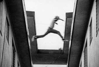 In a grainy black-and-white film still full of movement shot looking up at a roof from a small alleyway, a young male figure leaps from one roof to the next.