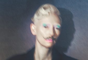 Op een enigszins onscherpe foto staat Tilda Swinton tegen een gevlekte grijze achtergrond, gekleed in een donker pak met dubbele rij knopen. Haar korte blonde haar is opgerold in een kuif, ze draagt een opvallende turquoise oogschaduw, een lichtroze lippenstift en een krullende bruine snor.