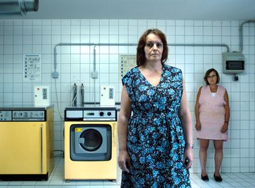 In een blauwachtige, betegelde kelderruimte staan ​​drie vrouwen rond twee gele wasmachines
