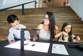 Een volwassene zit samen met twee kinderen achter de animatietafels in Eye, ze knippen vormen uit papier en plaatsen deze op een verlichte plaat om stop-motion animatiefilms op hun telefoon te maken