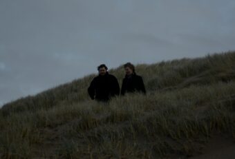 Foto van de twee muzikanten van Wanderwelle die door de duinen lopen, gekleed in zwarte jassen.