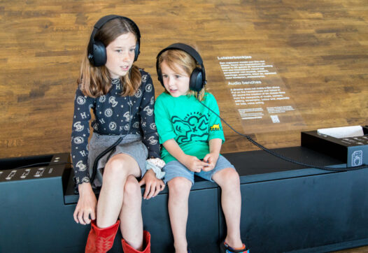 Twee kinderen luisteren met een hoofdtelefoon op naar verhalen op de Luisterbankjes