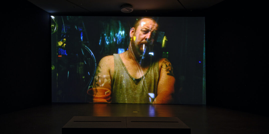 Beeld uit de tentoonstelling Cosmic Realism. Op een groot scherm zie je een stoere man die moeilijk kijkt met een sigaret in zijn mond.