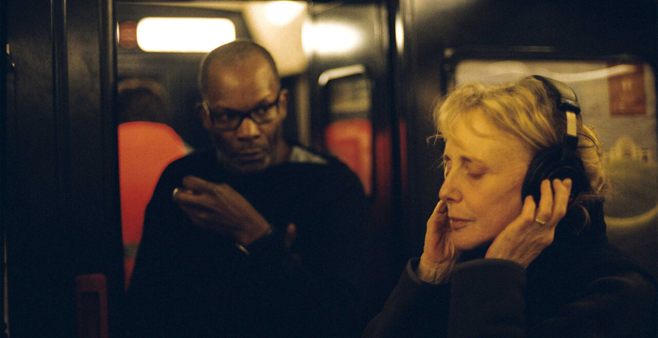 Op de voorgrond op een filmset staat Claire Denis, een vrouw met golvend blond haar, met haar handen op haar hoofdtelefoon, haar ogen gesloten terwijl ze aandachtig lijkt te luisteren. Op de achtergrond kijkt een man met een bril naar haar reactie