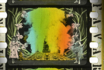 Een close-up van een gekleurde filmstrook met regenboogkleurige rookpluimen op de achtergrond, links en rechts staan twee figuren in sprookjesachtige jurken (campagnebeeld This is Film! 2023 (Het tovertoneel (Segundo de Chomón, FR 1907)))